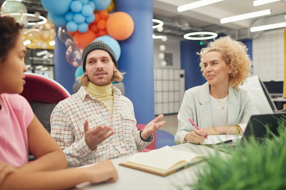Three young adults talking around a table in a colorful workspace, one gesturing as he explains an idea.
